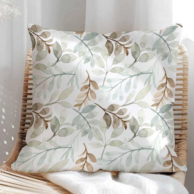 Almofada Folhas de Aquarela Verde Salvia e Marrom (Sage Green and brown watercolor leaves on a white throw pillow in a rattan armchair.)