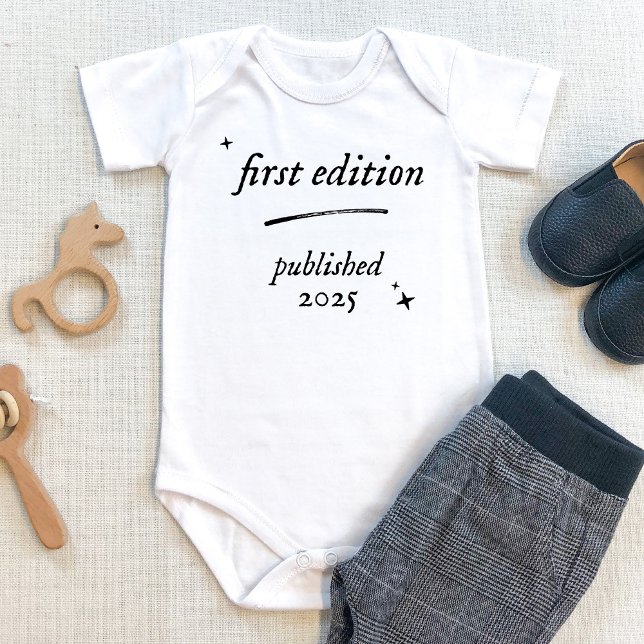 Body Para Bebê Livro da Primeira Edição Publicado em 2025 (First edition baby bodysuit with personalized birth year. Shown on beige table with baby pants. book)