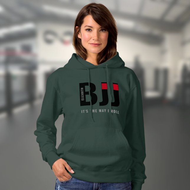 Moletom BJJ de mulher personalizado (Dark green woman's BJJ hoodie worn by a female model in a gym.)