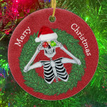 Ornamento De Cerâmica Esqueleto no Pine Christmas Wreath Red Hat Funny<br><div class="desc">Este ornamento cerâmico pendurado tem um esqueleto engraçado segurando ornamentos com flocos de neve até seus olhos vestindo um chapéu de Papai Noel vermelho dentro uma coroa verde sobre um fundo vermelho profundo. Personalize seu texto no verso com o e o Feliz Natal na frente com os modelos.</div>
