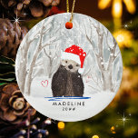 Ornamento De Cerâmica Ótila e fofa com o nome da Criança Santa Hat<br><div class="desc">Eterno e Enfeites de natal bonitinho com uma lontra adorável com um chapéu vermelho brilhante de Papai Noel. O fundo com tema aquarela apresenta uma sutil queda de neve invernal de pastéis mutados entre o cheio florestal de árvores. Dois corações vermelhos acompanham a lontra. A tipografia é um clássico sans...</div>