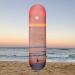 Rolando através do skate Sunset Ouro Laranja<br><div class="desc">Este skate mostra uma imagem de uma pessoa caminhando pelos raios ouro rosa e laranja do pôr do sol. Os raios coloridos criam uma órbita que silhueta o andarilho.</div>